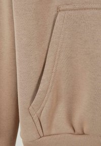 Hellbraunes Sweatshirt-Gewebe mit einer strukturierten Oberfläche, ausgestattet mit einer gesteppten Kängurutasche und einem gerippten Saum für zusätzliche Stabilität.
