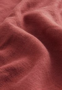 Tissu couleur rouille avec une surface douce et texturée, présentant des variations subtiles et des plis, mettant en valeur ses qualités légères et respirantes.
