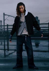 Giacca bomber nera sopra una camicia a quadri e una maglia a righe, abbinata a jeans scuri; presenta guanti senza dita e un dettaglio con portachiavi.