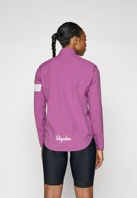Fioletowa kurtka rowerowa z wysokim kołnierzem, długimi rękawami, odblaskowymi białymi akcentami i zaokrąglonym dolnym wykończeniem, z logo "Rapha" z tyłu.