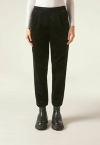 Pantalon en velours côtelé noir avec taille élastique, plis à l'avant, jambes fuselées, associé à des bottines noires. Tissu texturé avec des rayures verticales.