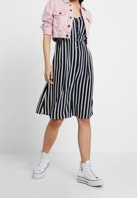 Abito a righe blu navy e bianche con scollo a camicia e gonna a campana, abbinato a un giubbotto di jeans rosa corto e sneakers bianche.