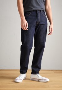 Tmavě modré džíny z denimu s rovně střiženými nohavicemi, kontrastním prošíváním, pěti kapsami a zapínáním na knoflíky vpředu, doplněné bílými teniskami.