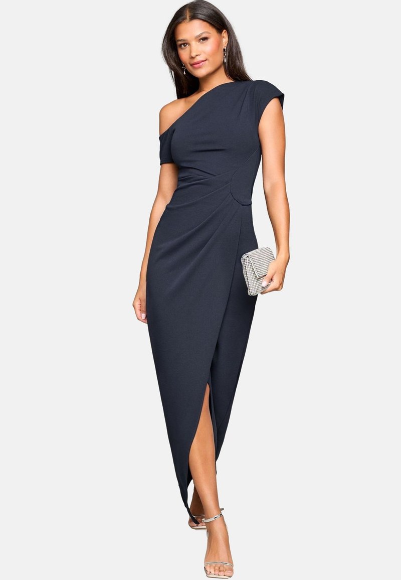 Donna che indossa un vestito nero monospalla con spacco frontale, tiene una pochette argento e indossa sandali argento con tacco alto.