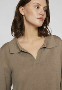 Top de punto marrón estilo polo con cuello y escote en V, que muestra una textura de canalé y un ajuste holgado. La tela parece suave y ligera.