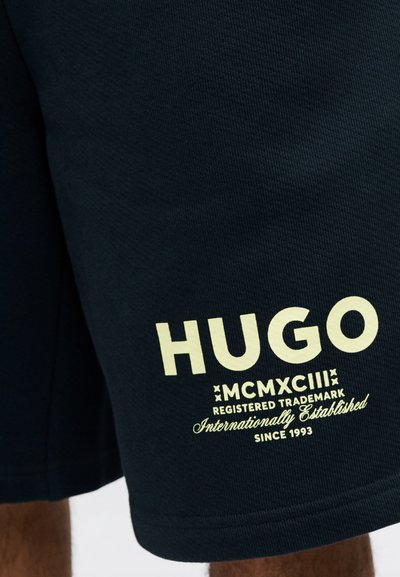 Крупен план на черен къс панталон с светъл надпис "HUGO MCMXCIII Регистрирана търговска марка Международно установена от 1993 г." на крачола.