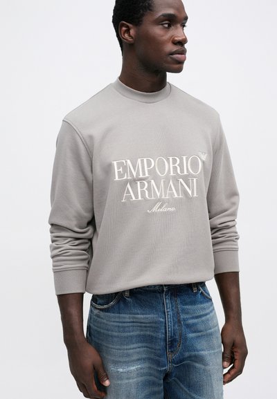 Sivá mikina s vysokým golierom, s bielym textom "Emporio Armani Milano". V kombinácii s modrými džínsami s jemným vyblednutím.