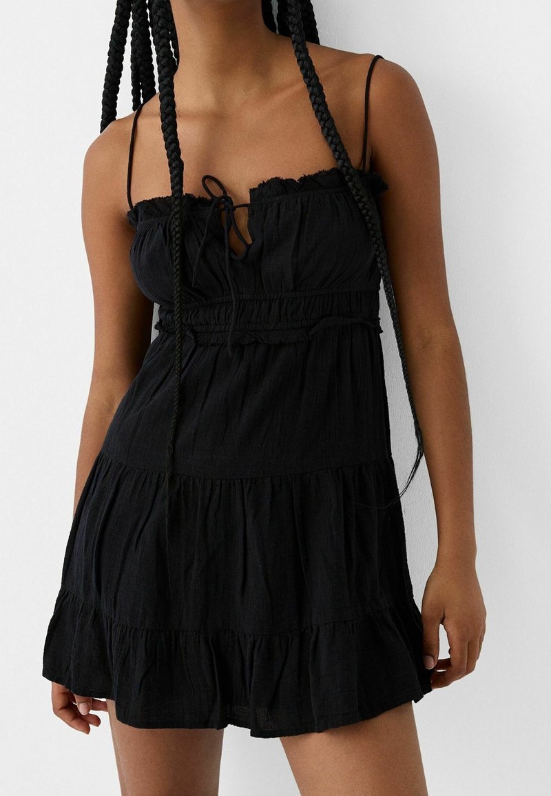 Svart sommarklänning med spaghetti-band, volangkrage, insydd midja och flerradig design. Tillverkad av ett lätt tyg med mjuk textur.