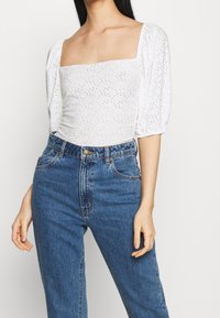 White puff-sleeve top with a square neckline, featuring eyelet detailing. Paired with high-waisted blue denim jeans.