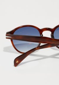 Lunettes de soleil rondes avec une monture en écaille de tortue marron et des verres dégradés bleus. Les branches texturées présentent un logo subtil près des embouts.