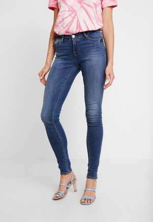 Vrouw draagt blauwe skinny jeans, roze tie-dye T-shirt met korte mouwen en lichtblauwe sandaal met hoge hakken, staand tegen een witte achtergrond.