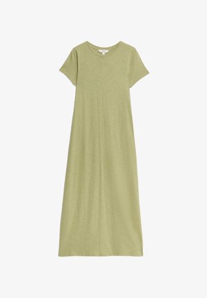 Personne portant une robe vert clair à manches courtes avec une veste blanche sans manches avec des poches, les mains reposant sur les hanches.