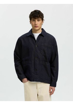 Ung mann som har på seg en mørk knappet jakke med tre lommer og lyse bukser, står foran en ensfarget bakgrunn.