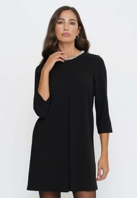 Schwarzes Shiftkleid mit Dreiviertelärmel, das einen runden Ausschnitt mit einer silbernen Perlenverzierung und einem geraden Schnitt aufweist.