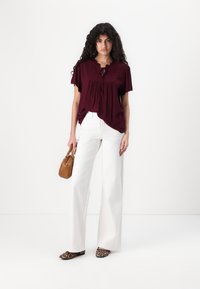 Blusa bordeaux con colletto arricciato e fiocco frontale, abbinata a pantaloni bianchi a gamba larga. La modella tiene una piccola borsa marrone e indossa scarpe con stampa leopardata.
