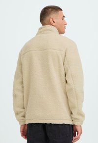 Beige fleece jas met een hoge kraag, gestructureerd oppervlak en een losse pasvorm. Heeft een eenvoudig ontwerp zonder zichtbare hardware of accenten.