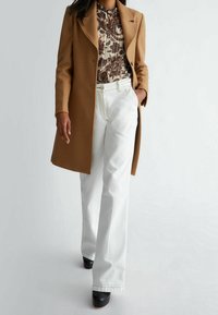 Brauner Wollmantel, weiße Jeans mit weitem Bein, gemustertes Blusentop und schwarze klobige High Heels. Glatte Stoffstrukturen und definierte Linien schaffen eine elegante Silhouette.