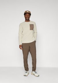 Homme portant un pull texturé beige avec une poche sur la poitrine, un pantalon marron, des baskets blanches et une casquette marron, se tenant devant un fond uni.