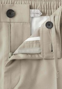 Pantalon beige avec braguette zippée ouverte, fermeture par bouton, taille élastique et boutonnières visibles.