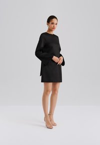 Svart minikjole laget av glatt stoff, med klokkeformede ermer, splitt bak og minimalistisk design. Kombinert med sølvfargede sandaler med remmer.