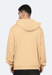 Felpa beige con cappuccio, maniche lunghe, polsini e orlo a costine. Texture morbida e vestibilità ampia, vista da dietro.