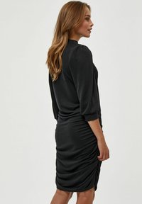 Crna haljina s dugim rukavima, nabrano detaljiranje u struku i prilagođenim krojem. Glatka tkanina pruža elegantan završetak.
