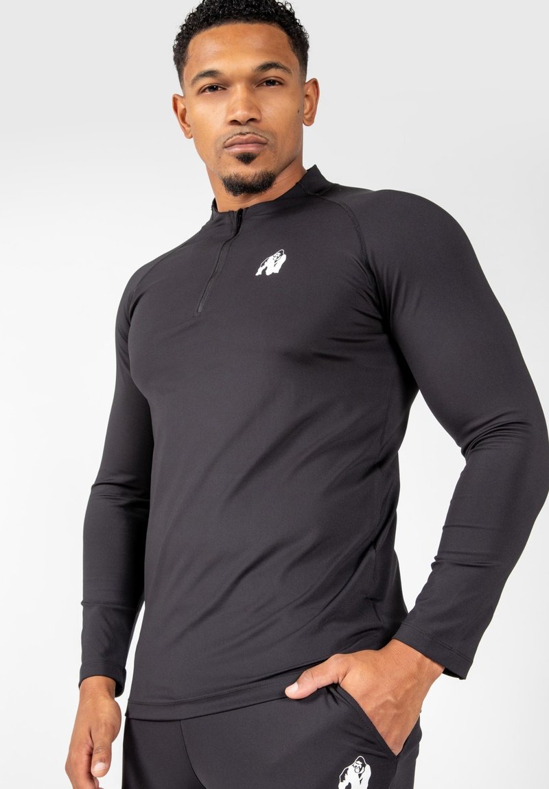 Zwarte sportieve langemouwenshirt met een kwart-rits kraag en wit logo op de borst. Glad, elastisch materiaal met een getailleerde pasvorm.