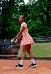 Tenisa spēlētāja gaiši oranžā īsās piedurknes krekliņā un svārkos, tur tennis racket un bumbu, ar melnajām kurpēm un baltām zeķēm uz māla korta.