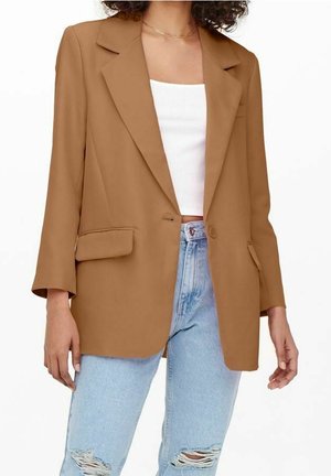 Kvinde iført en kamelfarvet blazer over en hvid cropped top og revnede lyseblå jeans, stående foran en hvid baggrund.