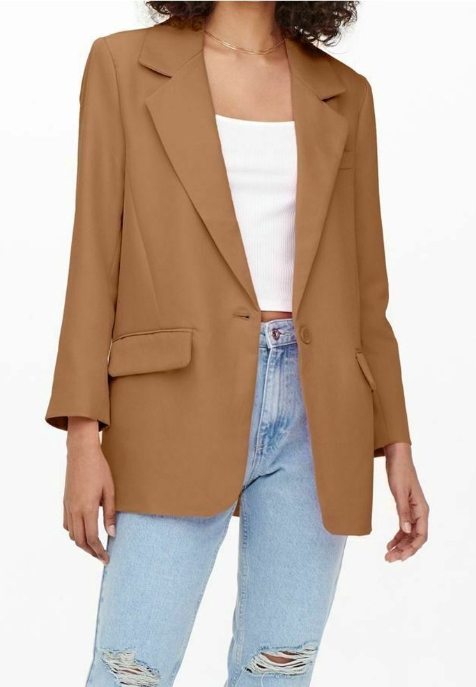 Donna con blazer color cammello sopra un top corto bianco e jeans strappati azzurri chiari, in piedi davanti a uno sfondo bianco.