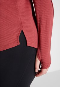 Langsleeve top in gestructureerd roestbruin, met een zijsplit, gestyled met zwarte broeken. De stof lijkt lichtgewicht en ademend te zijn.