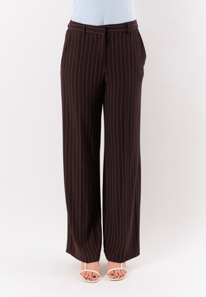 Donna che indossa pantaloni marroni a righe larghe e sandali bianchi, in piedi davanti a uno sfondo chiaro uniforme.