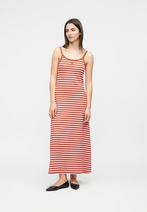 Femme portant une robe longue sans manches à rayures horizontales rouges et blanches, et des ballerines noires, debout devant un fond clair uni.