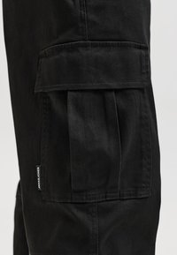 Gros plan sur la poche d’un pantalon cargo noir avec rabat et détail plissé, avec une petite étiquette de marque Jack & Jones cousue verticalement.