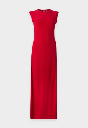 Mujer con cabello largo y rubio, vestida con un vestido rojo sin mangas hasta el suelo y tacones negros abiertos, de pie frente a un fondo claro y sencillo.
