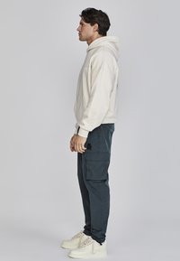 Cremefarbener Hoodie mit Seitentasche, kombiniert mit dunkelblaugrünen Cargo-Hosen mit aufgesetzter Tasche. Weiße Sneaker mit geflochtenen Schnürsenkeln runden den Look ab.