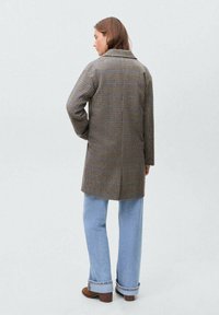 Rjav kariran plašč z udobnim krojem, ki ima zarezan ovratnik, zadnji razporek in ravno krojene rokave, v kombinaciji s svetlo modrimi kavbojkami.