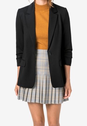 Donna che indossa un blazer nero sopra un top senape a coste abbinato a una minigonna plissettata grigia a quadri, in piedi davanti a uno sfondo bianco.