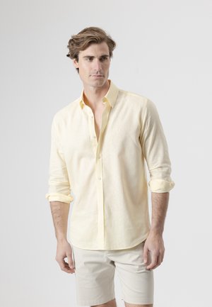 Joven con las mangas remangadas, vistiendo una camisa amarilla clara abotonada y pantalones cortos beige, de pie frente a un fondo gris liso.