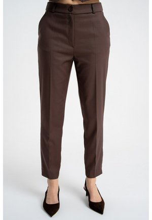 Kobieta w brązowych spodniach z wysokim stanem i prostą nogawką oraz w brązowych szpilkach z ostrym czubkiem, stojąca na tle jednolitego, jasnego tła.