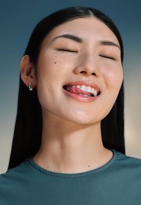 Close-up van een persoon met een gladde huid en lang, donker haar, gekleed in een teal top. Opvallende kenmerken zijn de gesloten ogen en een lichte glimlach die tanden onthult.