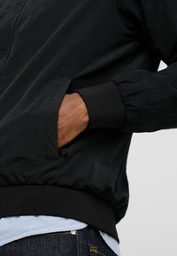 Veste noire légère avec des poignets élastiques, un détail de poche et un tissu texturé. Présente un discret patch logo sur le côté.