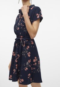 Marinblå blommig klänning med korta ärmar, insvängd midja och matchande bälte. Har rosa blommor och en avslappnad kjol. Slät tygtextur.