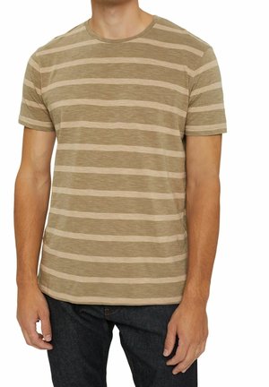 Dorosła osoba ubrana w krótki rękaw w poziome beżowo-brązowe paski i ciemnoniebieskie dżinsy, stojąca z ramionami swobodnie opuszczonymi wzdłuż ciała.