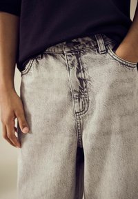 Graue Jeans aus Denim mit strukturiertem Finish, hoch geschnitten, mit Fronttaschen und dezenten Verblassungen, von einer Hand in der Nähe des Taillenbunds gehalten.