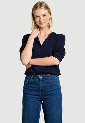 Frau mit kurzen blonden Haaren, die goldene Creolen, eine marineblaue Strickjacke und blaue Jeans trägt, posiert mit einem verschränkten Arm und einer Hand nahe am Gesicht.