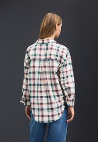 Camicia di flanella con bottoni, base bianca e motivo a quadri verdi, rosa e rossi. Presenta maniche lunghe e orlo curvo. Tessuto texturizzato.