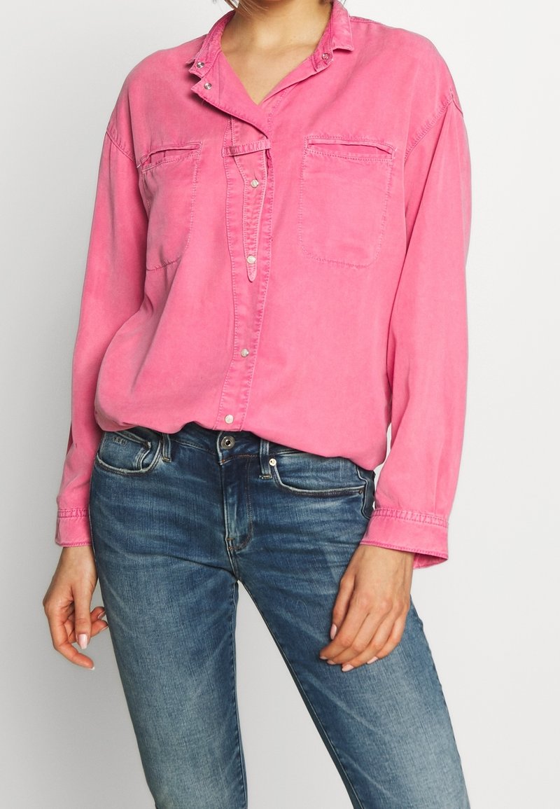 Personne portant une chemise rose boutonnée avec des poches poitrine et un jean en denim bleu sur un fond clair uni.