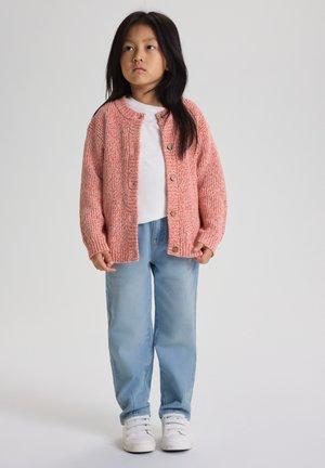 Kind staat en draagt een koraalkleurige gebreide cardigan over een wit shirt, lichtblauwe spijkerbroek en witte sneakers tegen een effen achtergrond.