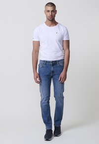 Bela majica s kratkimi Rokavi in majhnim logotipom, v kombinaciji z modrimi slim-fit jeansi in temnimi supergami, prikazana na nevtralnem ozadju.